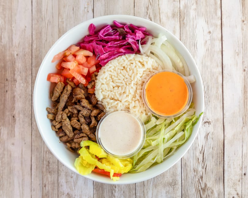 A white bowl filled with rice, meat, tomatoes, cabbage, onions, and two sauces on a wooden table.