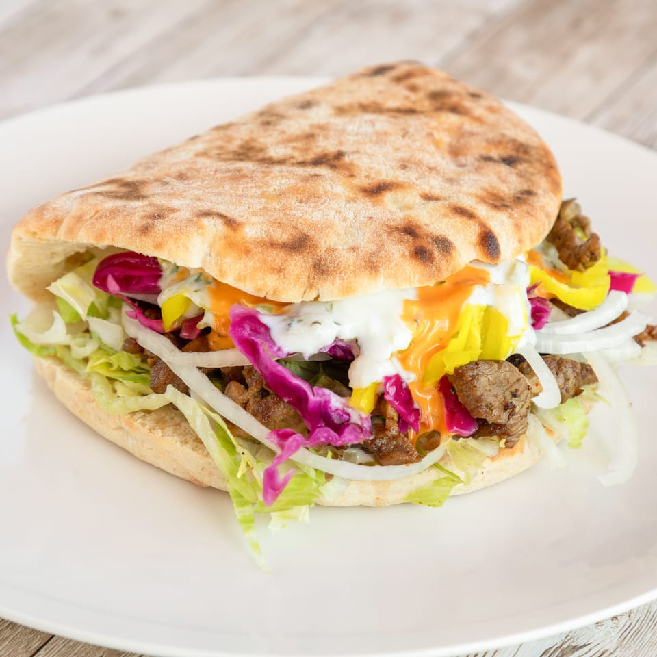 A pita bread sandwich filled with meat, lettuce, red cabbage, onions, and sauces on a white plate.