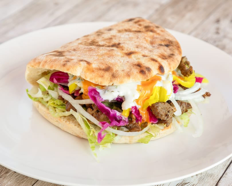 A pita bread sandwich filled with meat, lettuce, red cabbage, onions, and sauces on a white plate.