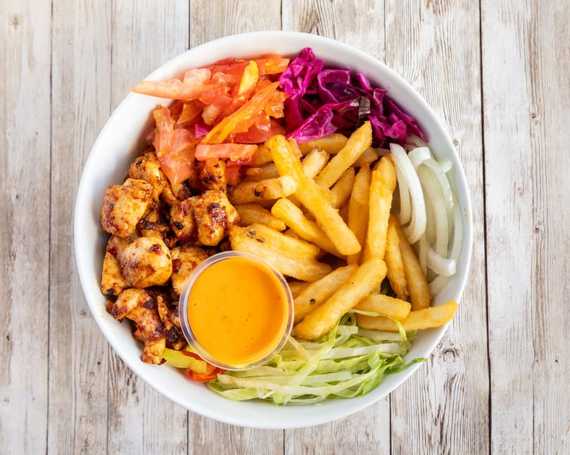A white bowl filled with grilled chicken, french fries, red cabbage, tomatoes, onions, lettuce, and sauce.