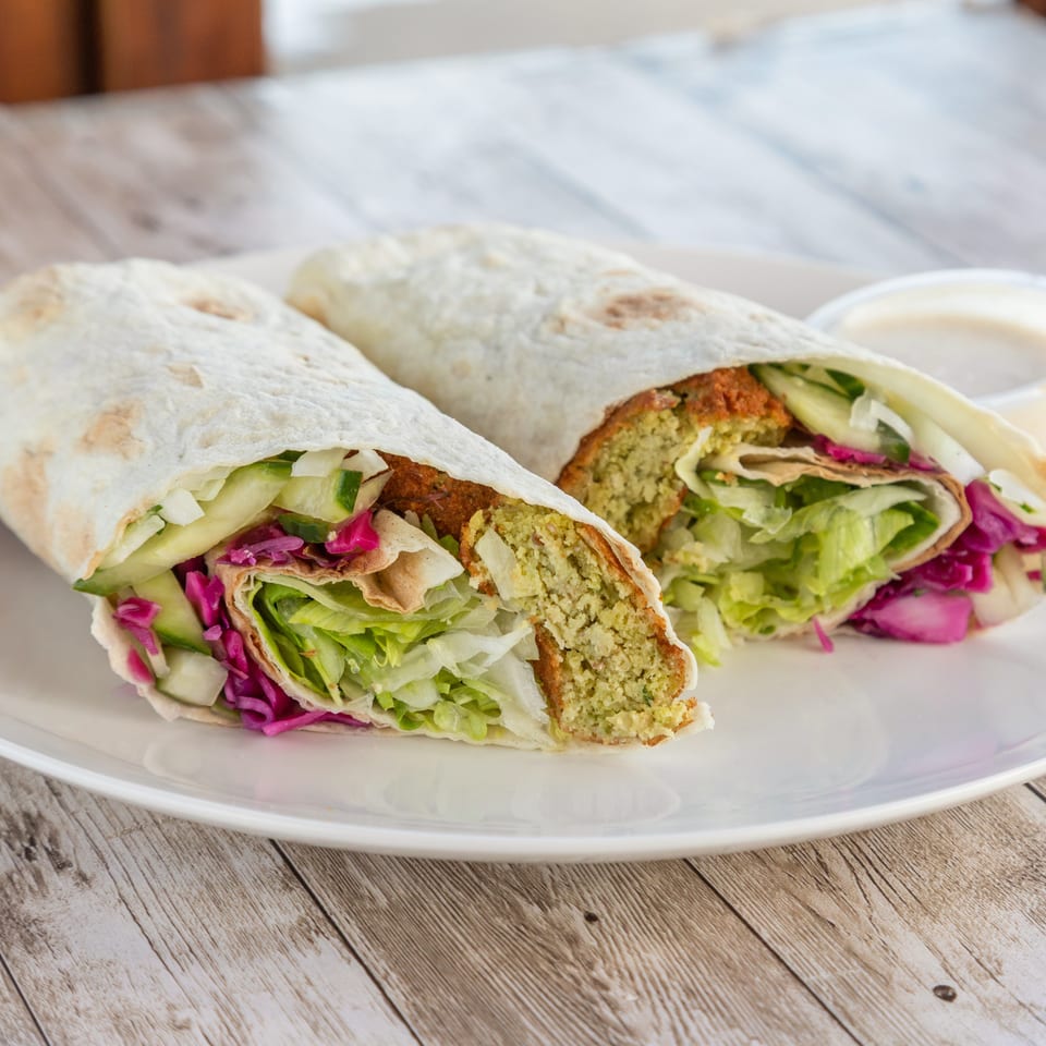 Two falafel wraps, cut in half, filled with lettuce, cucumber, and red cabbage, with a side of sauce.