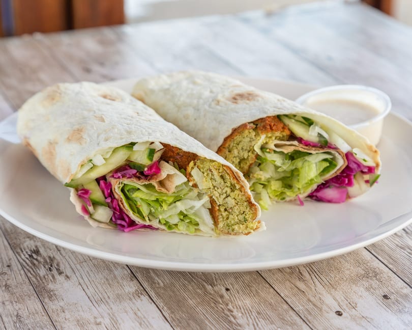 Two falafel wraps, cut in half, filled with lettuce, cucumber, and red cabbage, with a side of sauce.