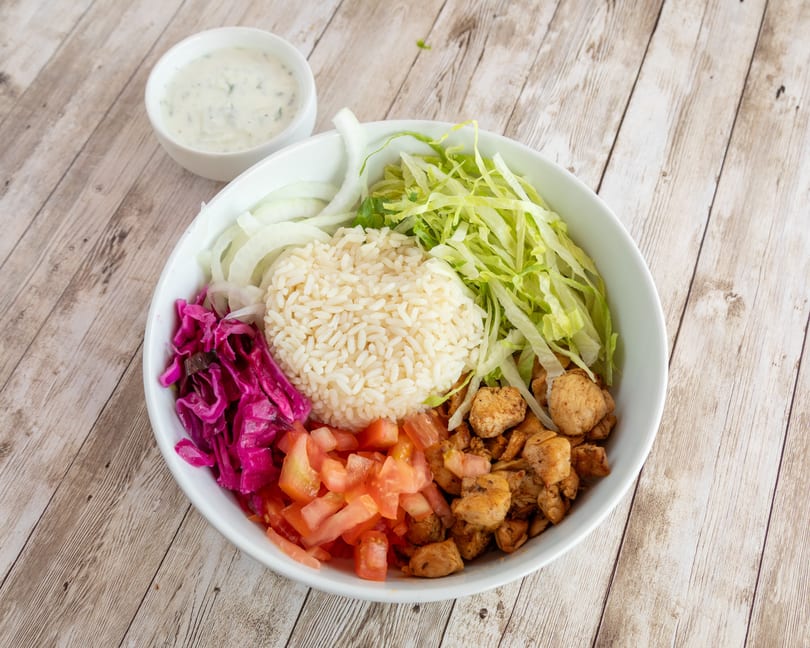 Chicken rice bowl with lettuce, onions, red cabbage, tomatoes, and a side of sauce.