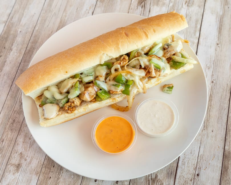 A chicken cheesesteak sandwich with melted cheese, green peppers, and onions, served with two dipping sauces.