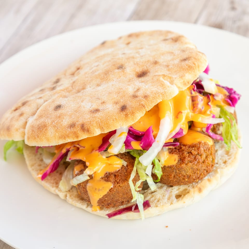 Falafel pita sandwich with lettuce, red cabbage, onions, and orange sauce on a white plate.