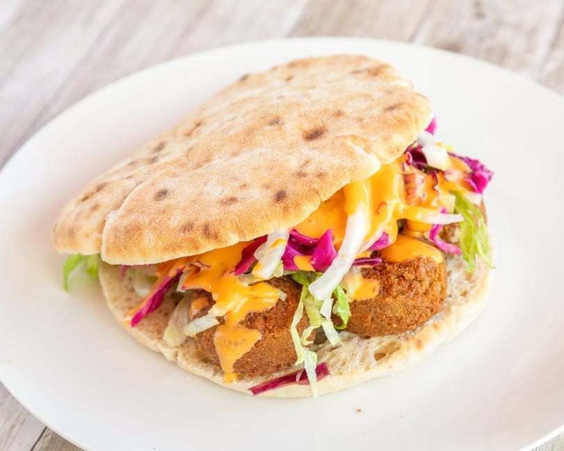 Falafel pita sandwich with lettuce, red cabbage, onions, and orange sauce on a white plate.