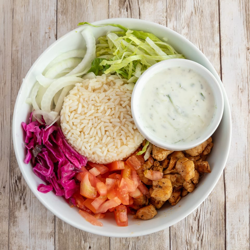A white bowl filled with rice, chicken, tomatoes, red cabbage, onions, lettuce, and a side of sauce.