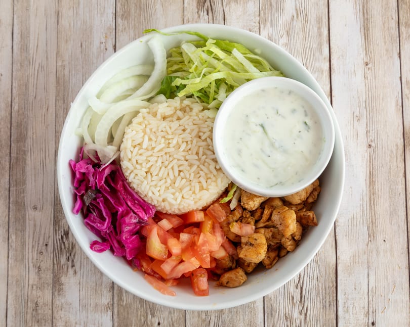 A white bowl filled with rice, chicken, tomatoes, red cabbage, onions, lettuce, and a side of sauce.