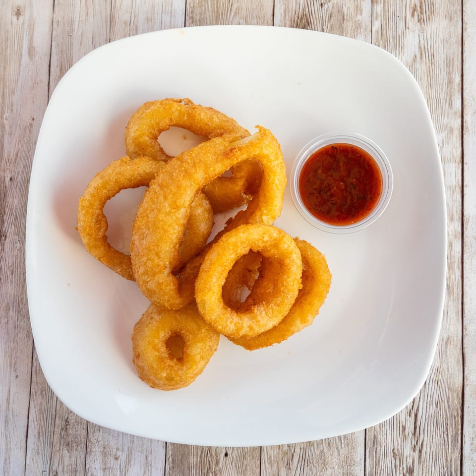 Golden-brown onion rings piled on a white plate with a side of red dipping sauce.