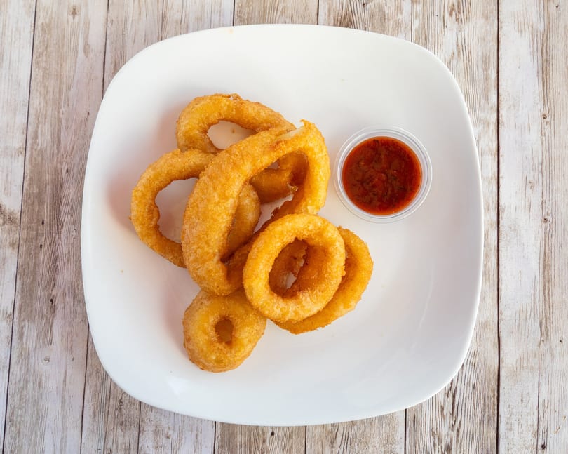 Golden-brown onion rings piled on a white plate with a side of red dipping sauce.