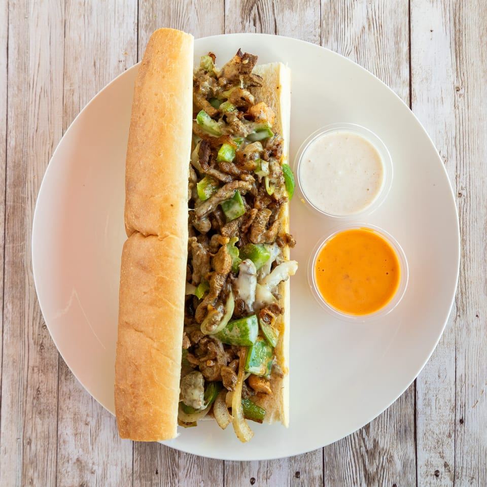 Cheesesteak sandwich with green peppers and onions, served with two dipping sauces on a white plate.