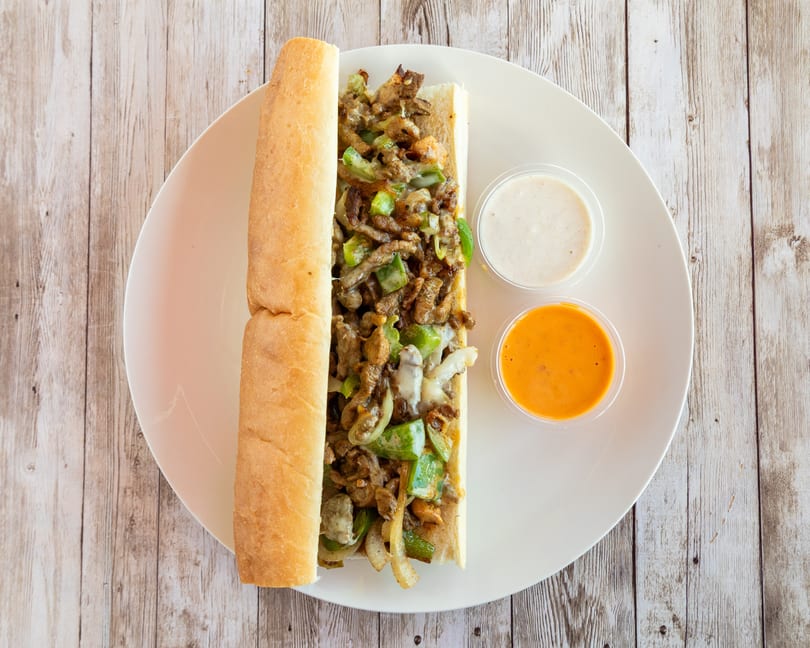 Cheesesteak sandwich with green peppers and onions, served with two dipping sauces on a white plate.